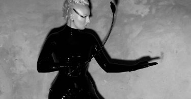black and white image of blonde femme person wearing latex and holding crop - is she a domme or dominatrix