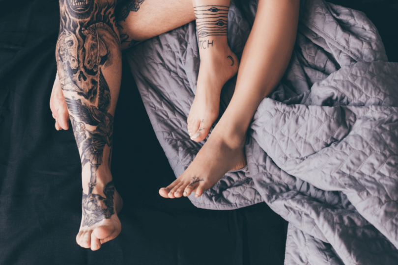 tattoed legs of man and woman in bed with rumpled blankets