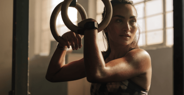 woman in gym with hair pulled back, holding onto rings, arm muscles flexed as concept for central character in erotic fantasy