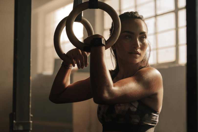 woman in gym with hair pulled back, holding onto rings, arm muscles flexed as concept for central character in erotic fantasy