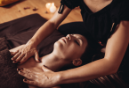 woman lying down on floor with towel covering chest and eyes closed as feminine hands massage her chest with a candle in the background as a concept for tantric massage