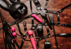 wood table with hot pink vibrators and black leather fetish gear including a mask, a crop, a flogger, and a harness