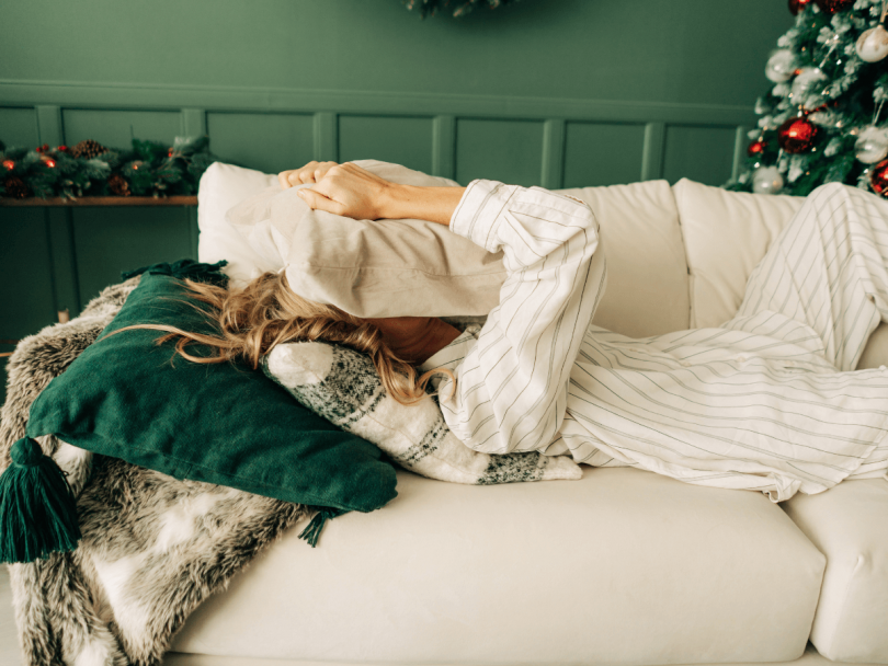 person in pajamas lying on couch with pillow over face and Christmas tree in the background as a concept for being stressed during the holidays